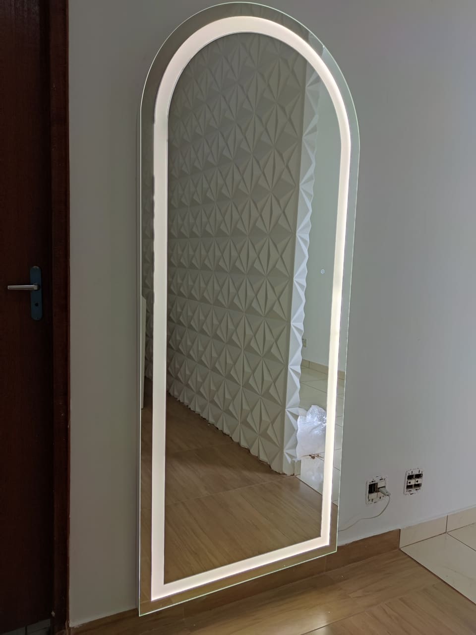 Espelho em arco de corpo inteiro com LED touch aceso ao lado de porta de madeira, refletindo parede 3D branca