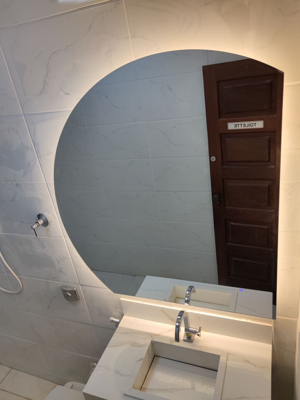 Espelho orgânico com LED quente em lavabo de porcelanato claro, refletindo a porta de madeira