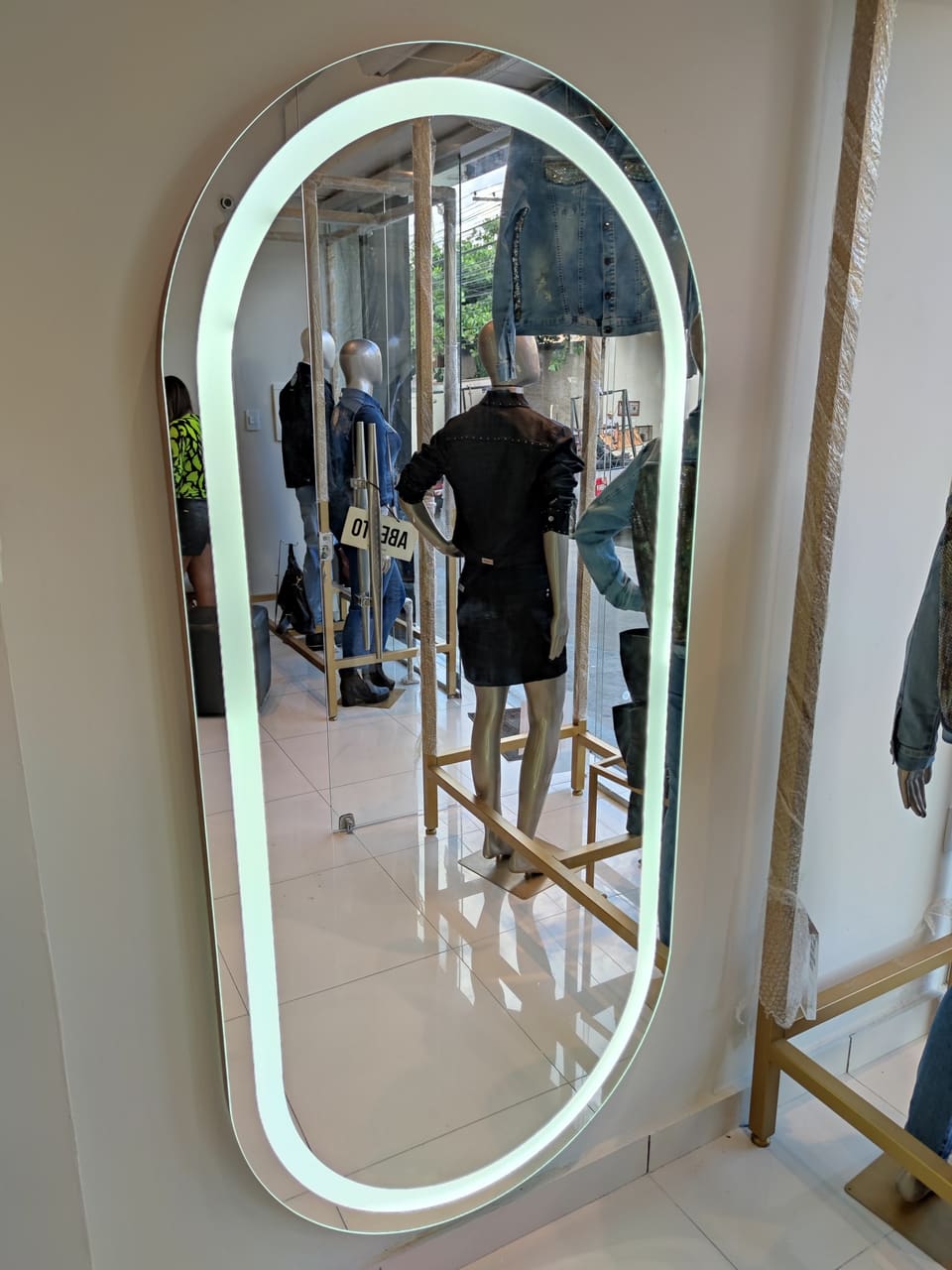 Espelho oval formato cápsula com LED frontal em loja de roupas, refletindo manequins